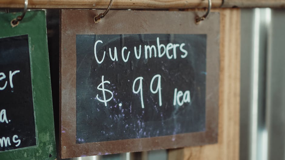 Close-up of a blackboard-style price sign displayed on a wooden surface at a gardening market, indicating that cucumbers are sold at $0.99 each. The sign features handwritten white chalk text, with th